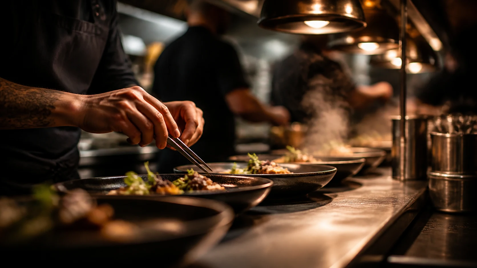 Open kitchen pass with chef hands plating dishes under warm heat lamps